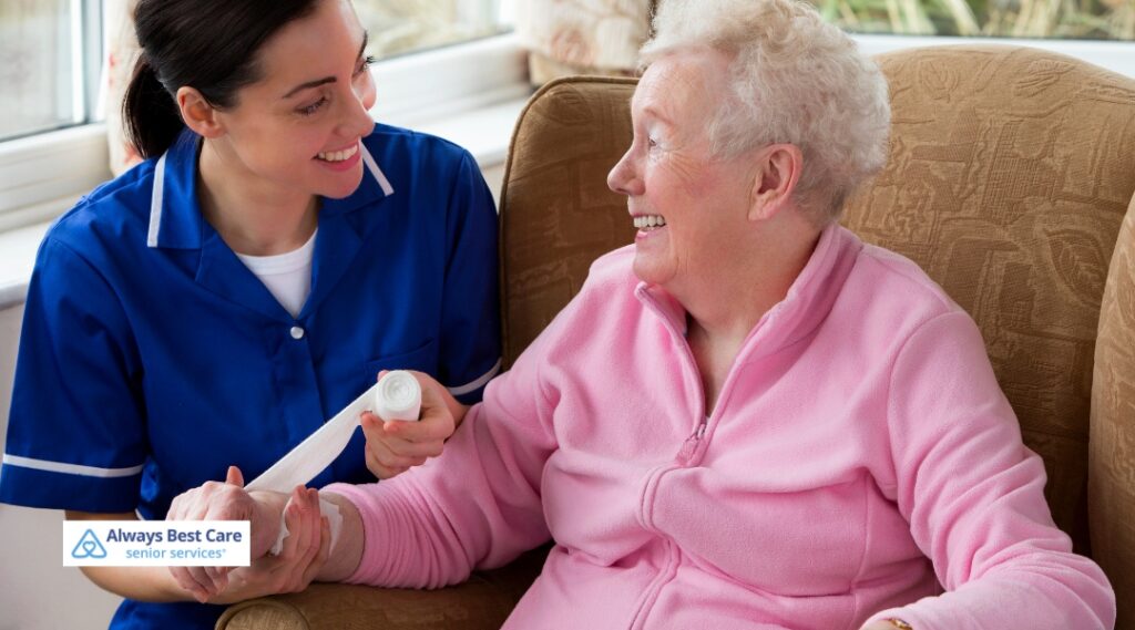 Nurse in blue uniform wrapping a bandage around an elderly woman's hand while sharing a warm smile in a cozy home living room.