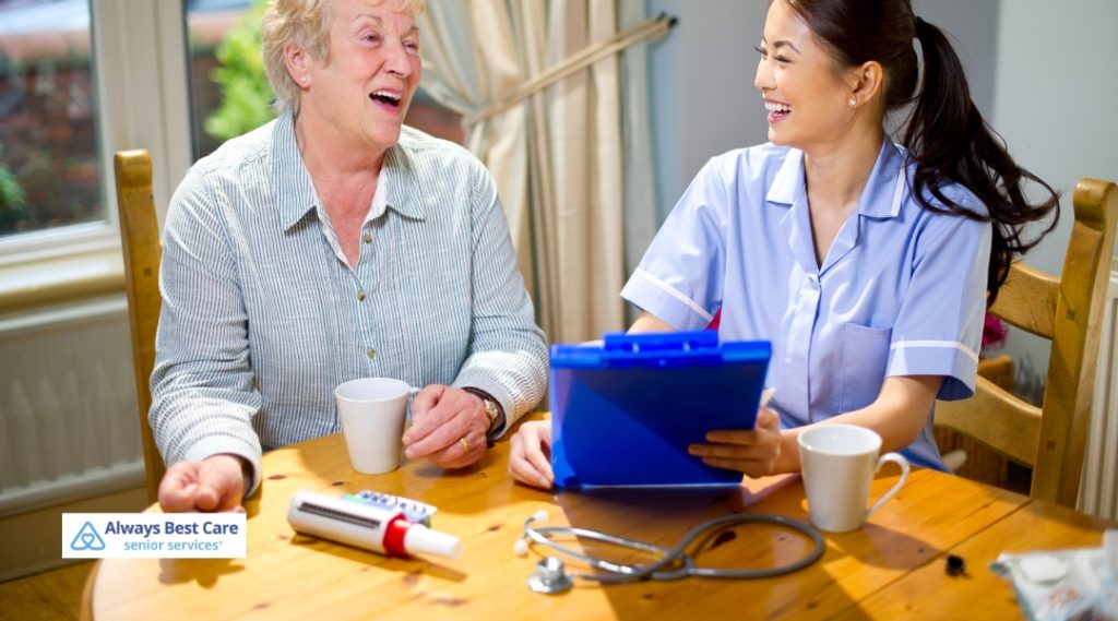 A caregiver in a blue uniform sitting next to an elderly man, smiling and reading from a book together while enjoying a cup of coffee.