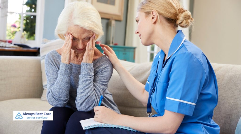 A caregiver in a blue uniform comforting an elderly woman sitting on a sofa, who appears to be distressed and holding her head.