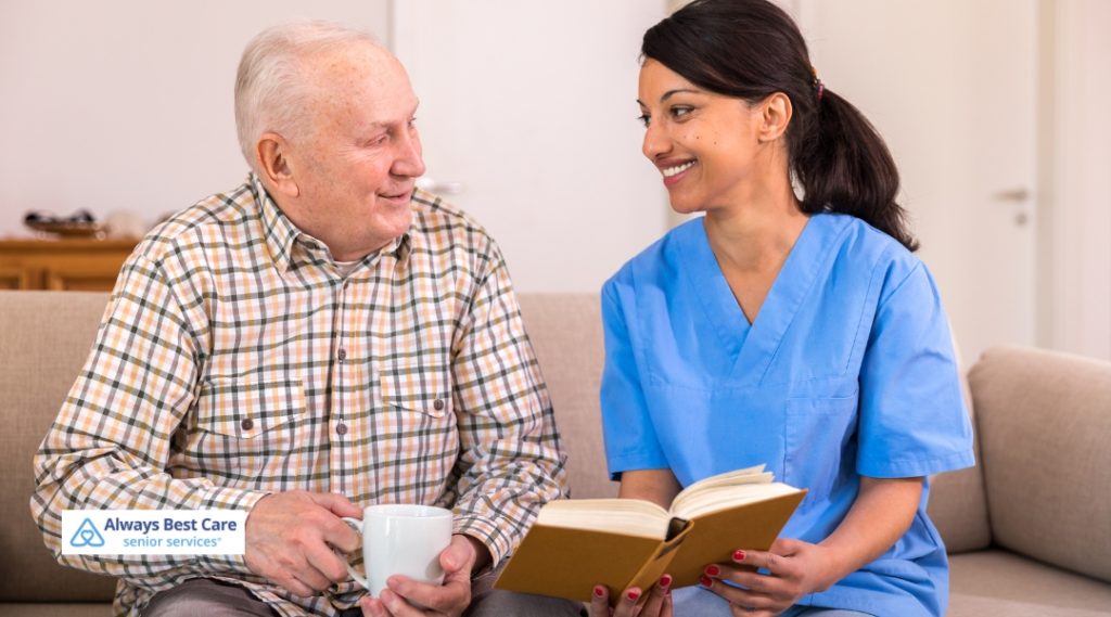 A caregiver in a blue uniform sitting next to an elderly man, smiling and reading from a book together while enjoying a cup of coffee.
