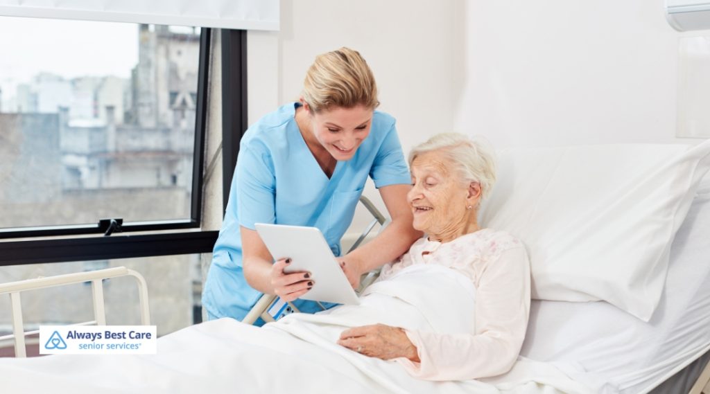A caregiver in a light blue uniform assisting an elderly woman in a hospital bed, showing her something on a tablet while both smile.