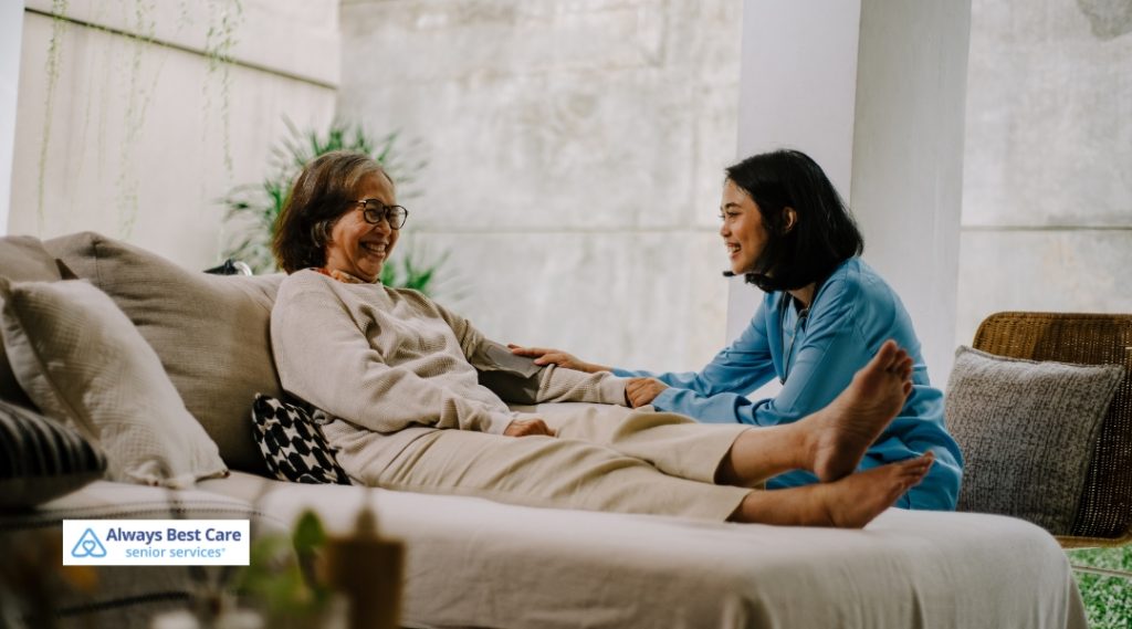 A caregiver in a light blue uniform providing assistance to an elderly woman resting on a sofa, with both smiling and engaging in conversation.
