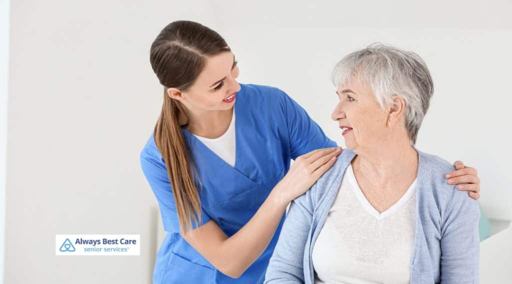A young female caregiver gently places her hands on the shoulders of a smiling senior woman, sharing a warm, encouraging interaction in a clinical or caregiving environment.