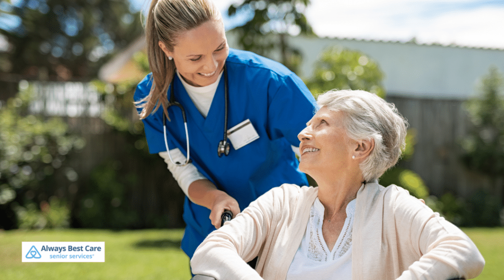 A healthcare professional in blue scrubs talks to an elderly woman sitting in a wheelchair outdoors in a sunny garden, both making eye contact and smiling.