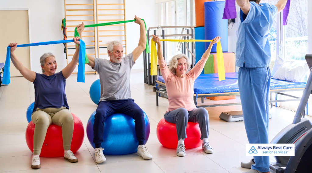 Three older adults sit on large exercise balls and raise colorful resistance bands overhead during a group exercise session while a trainer in blue scrubs leads them in a bright rehab gym; ‘Always Best Care senior services’ logo in the corner.