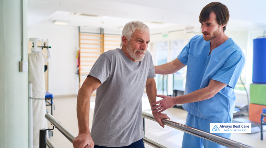 Older man practices walking while holding parallel bars as a caregiver in blue scrubs supports him in a bright rehab room; ‘Always Best Care senior services’ logo in the corner