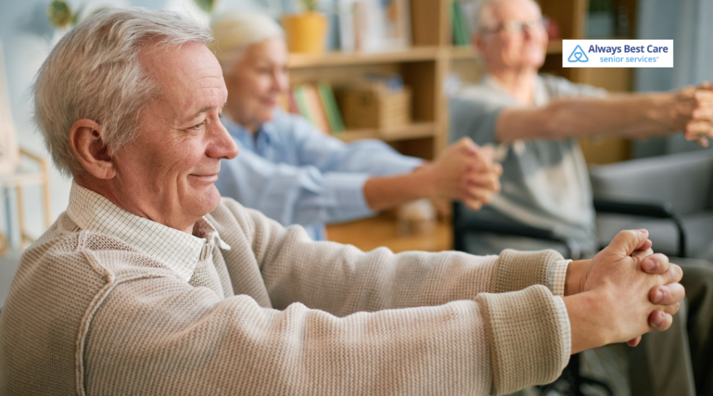 Older adults do seated stretching with hands clasped and arms extended in a cozy room; one man smiles in the foreground; ‘Always Best Care senior services’ logo in the corner.
