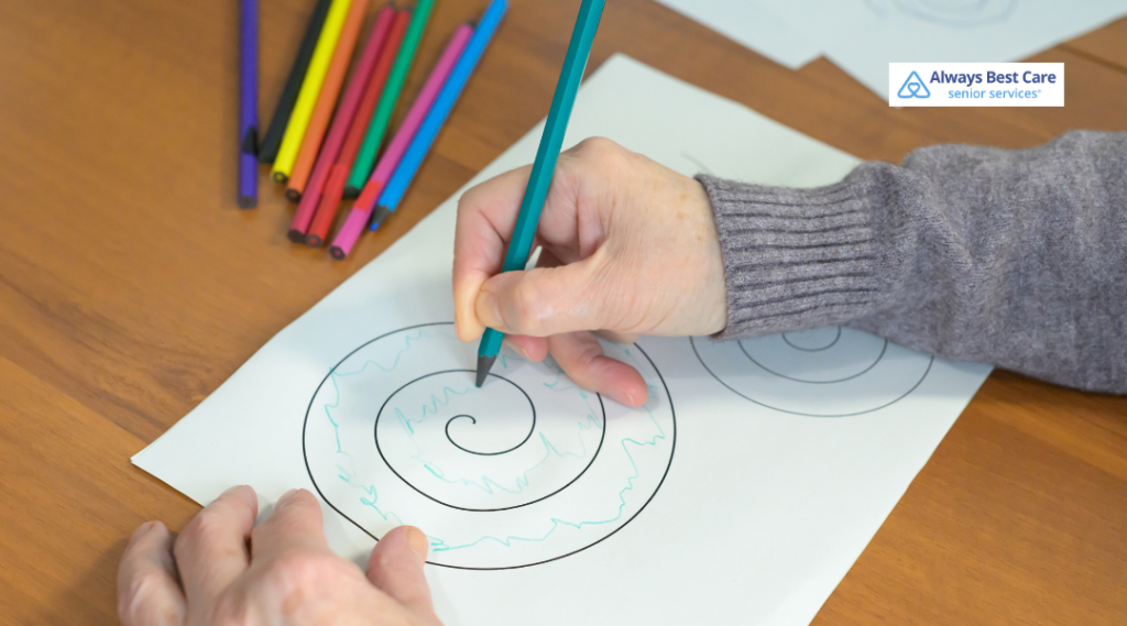 Close-up of hands drawing a spiral pattern on white paper with a green pencil; colored markers on a wooden table; ‘Always Best Care senior services’ logo in the corner.