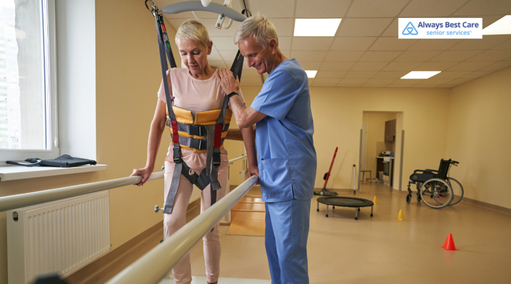 Senior woman in a support harness holds parallel bars while a therapist in blue scrubs guides her during gait training.