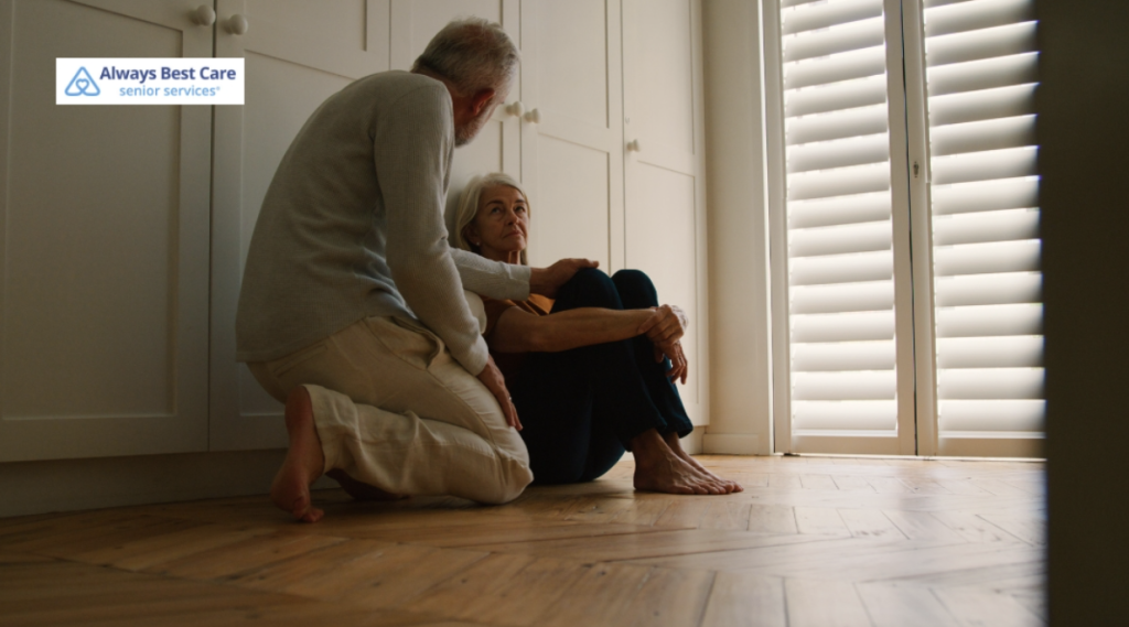 A senior woman sitting on the floor with a concerned expression, while a senior man kneels next to her, offering comfort and support. The room features soft, natural light coming through shutters.