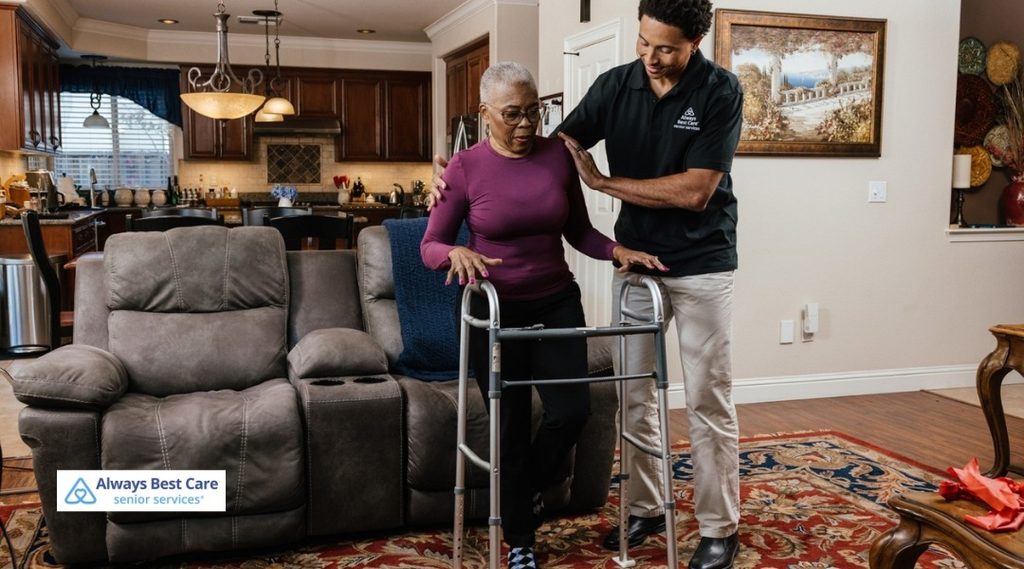 Caregiver supporting a senior woman as she uses a walker in her living room.