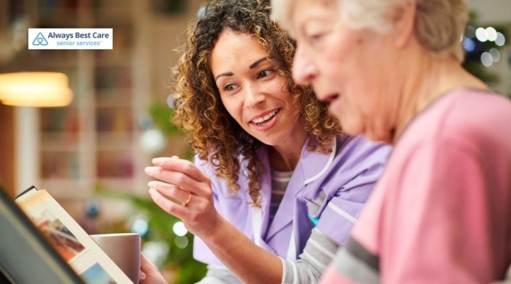 A caregiver with curly hair and a light purple uniform smiles warmly while helping an elderly woman in a pink sweater look at a photo album or book. The setting is cozy and home-like.