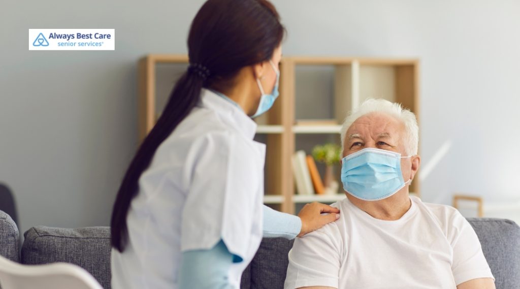 A caregiver, wearing a face mask and a white medical uniform, sits beside an elderly man who is also wearing a face mask. The caregiver has her hand gently resting on his shoulder as they engage in a conversation.