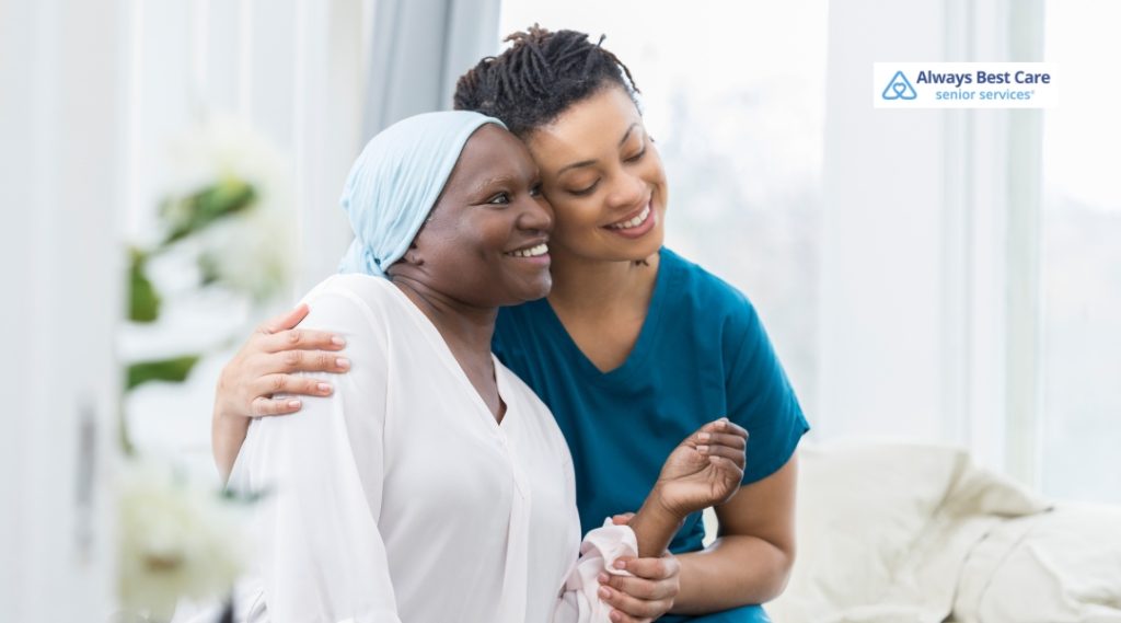 A caregiver, in teal scrubs, shares a joyful moment with an elderly woman wearing a light blue headscarf. The two are seated close together, smiling and embracing in a cozy indoor setting.