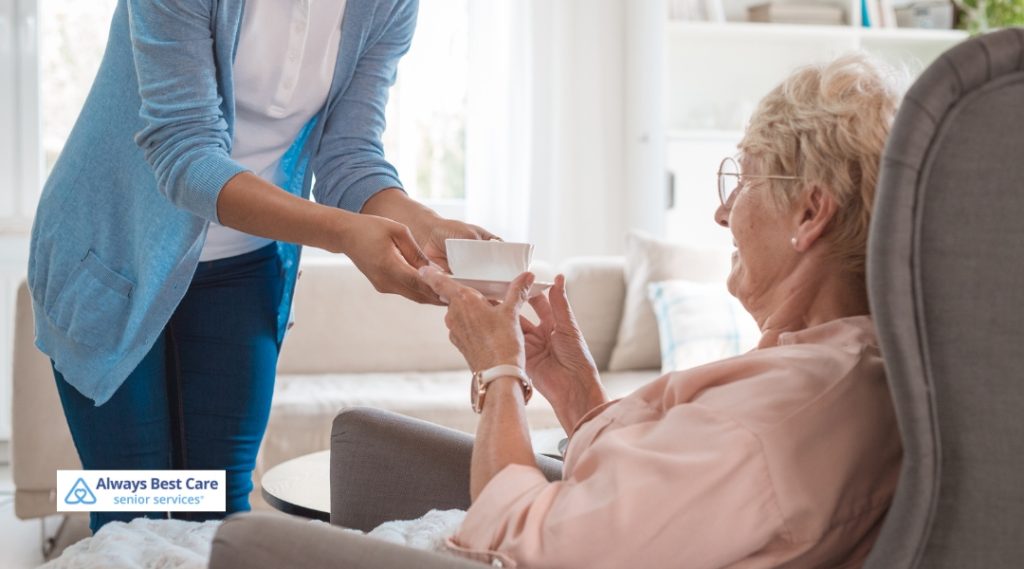 A caregiver, dressed in a light blue sweater, hands a bowl to an elderly woman sitting comfortably in a chair. The elderly woman, wearing a light pink shirt, reaches out to receive the bowl, with a look of appreciation on her face.