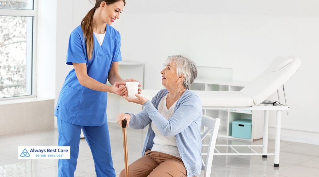 A caregiver in bright blue scrubs hands a white cup to a seated elderly woman with a cane, both smiling in a clean, clinical setting.