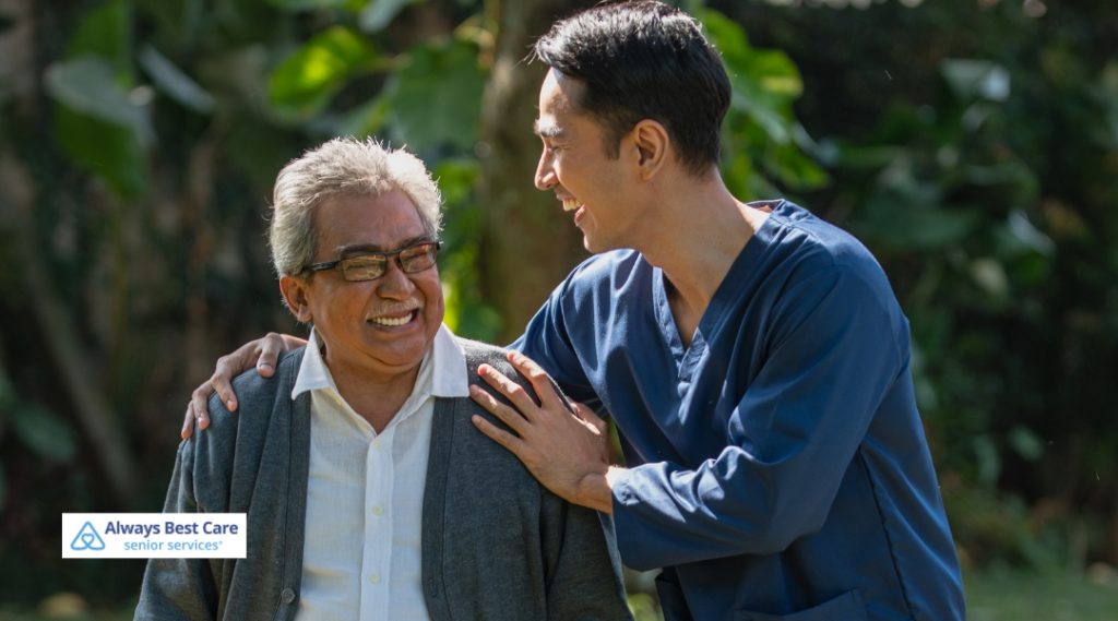 A young male caregiver in navy scrubs chats with and supports an elderly man wearing glasses and a cardigan, both smiling outdoors among greenery.
