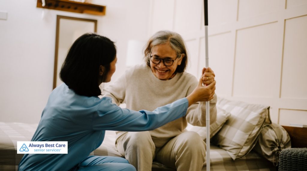 A female caregiver in scrubs helps a seated senior woman holding a white cane, both smiling warmly in a bedroom with natural lighting.