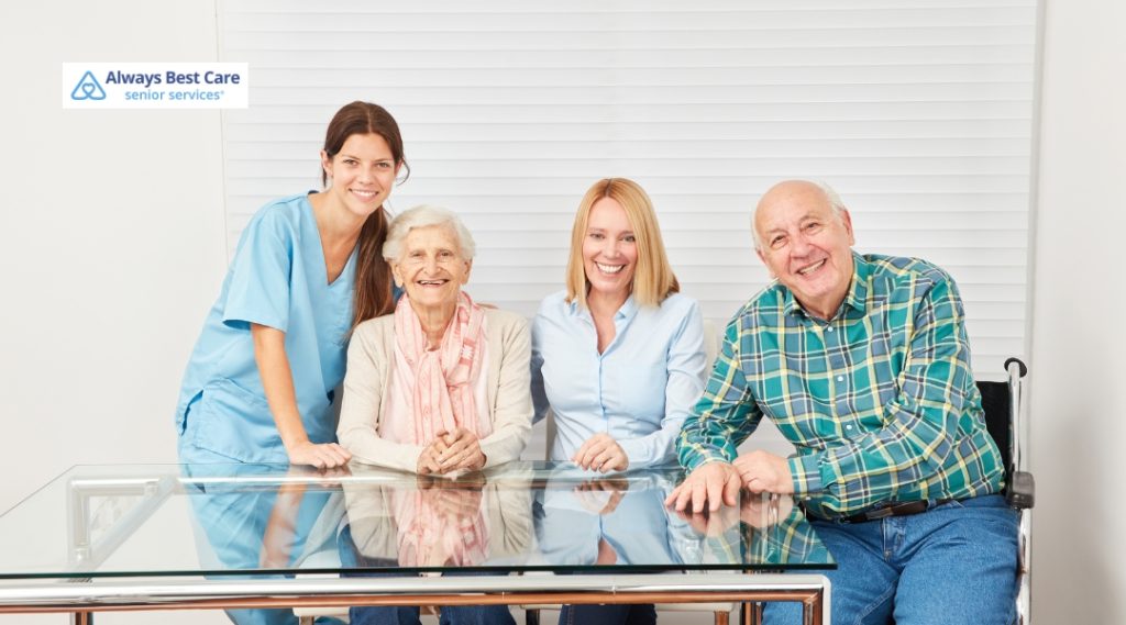 A caregiver in medical scrubs poses with two elderly individuals, one seated at a table and the other in a wheelchair. The scene takes place indoors with a light-colored backdrop and modern decor.