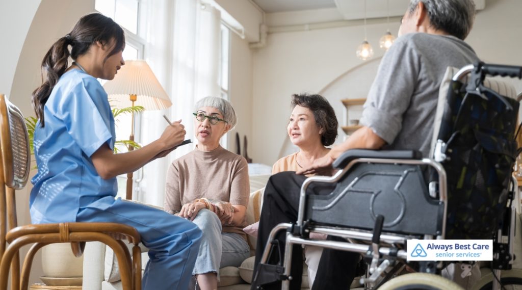 A caregiver in blue scrubs talks with a small group of elderly individuals, two of whom are seated in wheelchairs. The scene shows a peaceful indoor environment with natural light streaming in from windows.