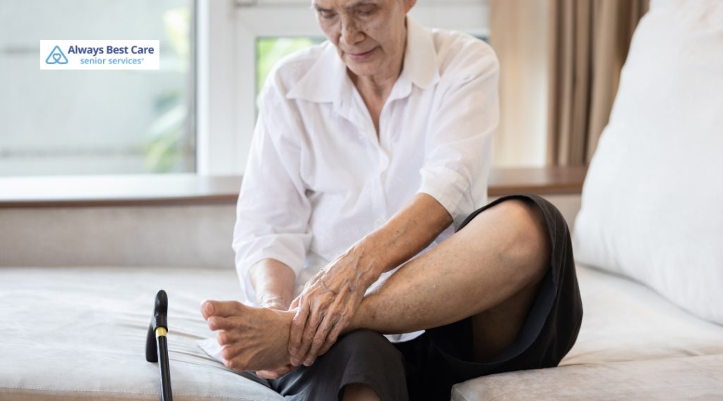 A senior person sitting on a sofa, dressed in a white shirt and dark skirt, grasps their ankle with both hands for support. 