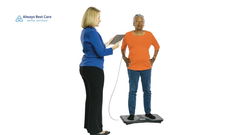 An older adult stands on an electronic monitoring device with hands on hips while a professional in blue attire uses a tablet to record health data during a wellness check. 