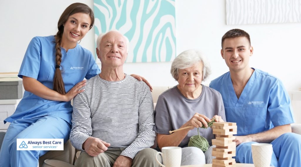 Two caregivers and two seniors engage in a fun and interactive activity at home. One caregiver stands beside a man, while the other sits beside a woman, both of whom are playing a game together. The atmosphere is relaxed and joyful.