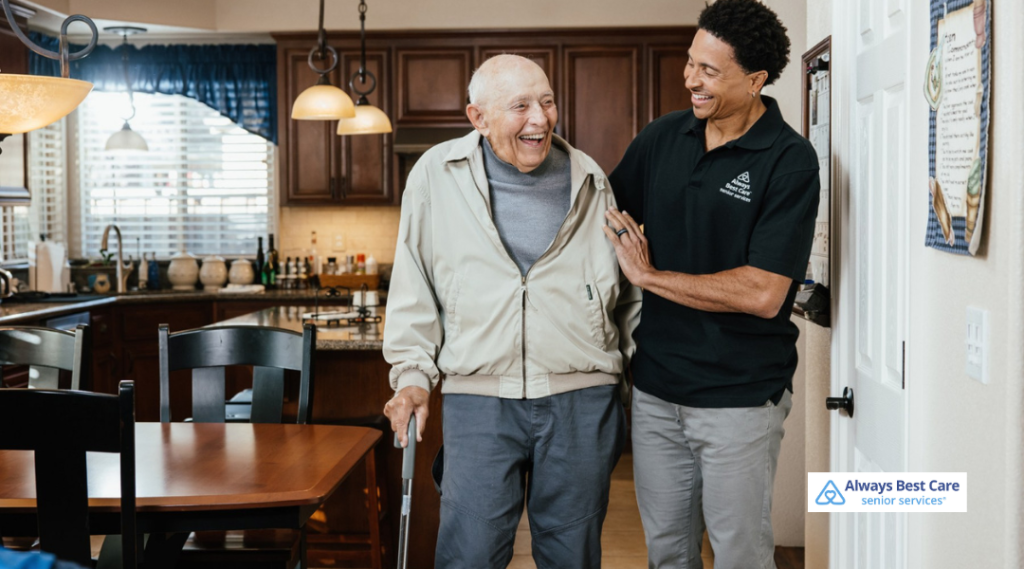Caregiver supporting an elderly man with a cane as they walk safely through his home.