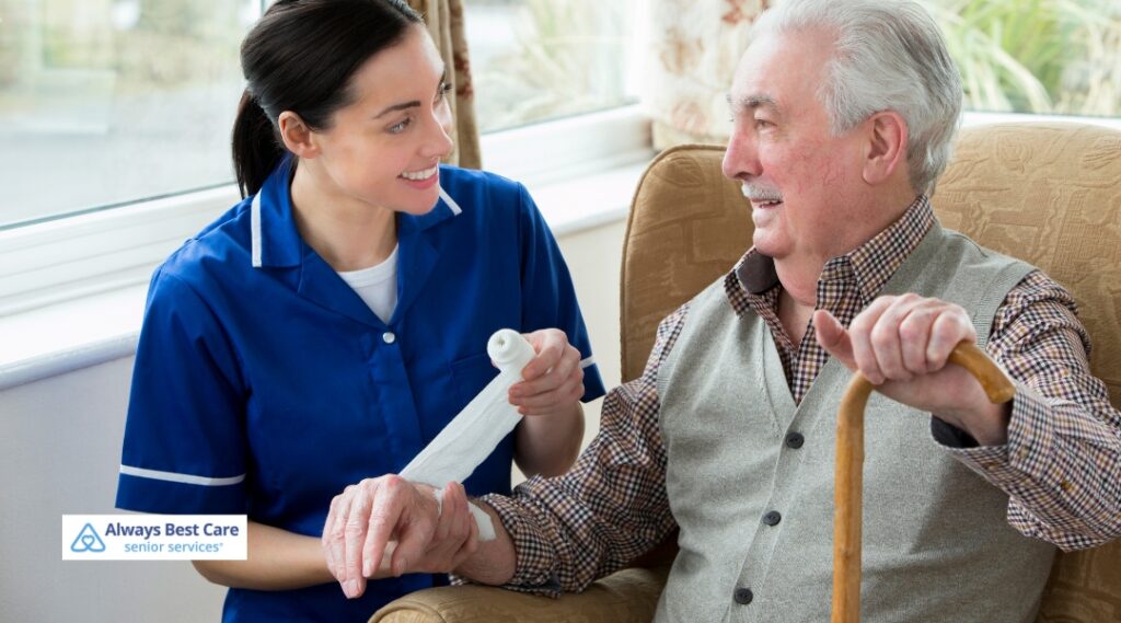 Skilled nurse bandaging the hand of an elderly man with a walking cane, both smiling during an in-home wound care session.