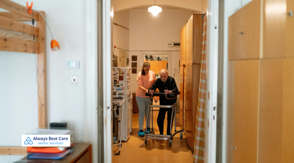 Caregiver walking beside an older man using a walker in a hallway at home, showing mobility assistance and daily support from Always Best Care.