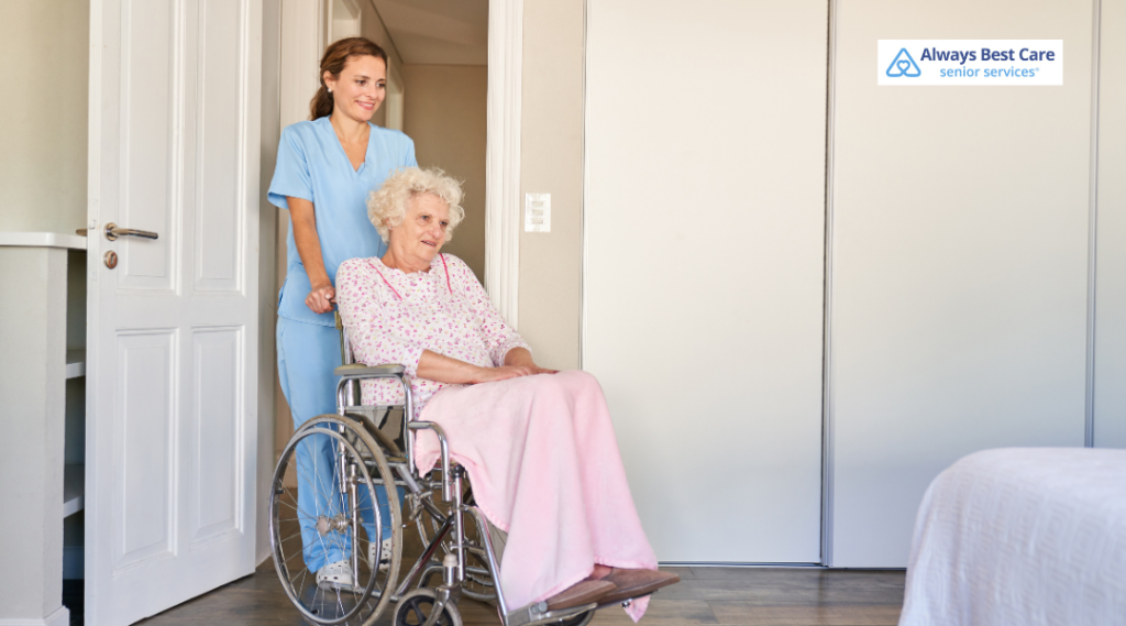 A professional caregiver in blue scrubs assists an elderly woman in a wheelchair, guiding her through a doorway in a comfortable home setting.