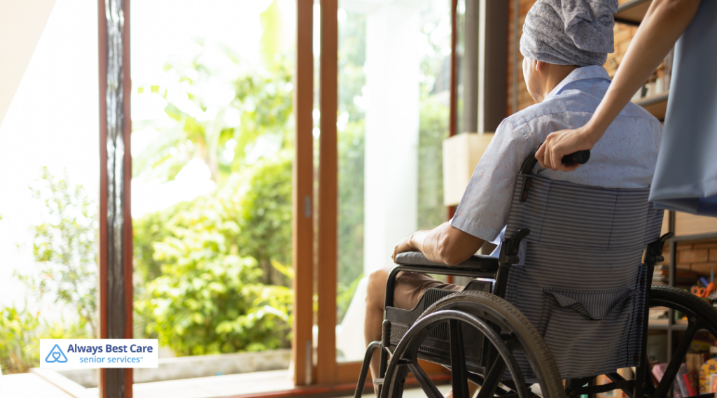 An elderly person wearing a headscarf is seated in a wheelchair near a sunlit open doorway, being gently guided by a caregiver.