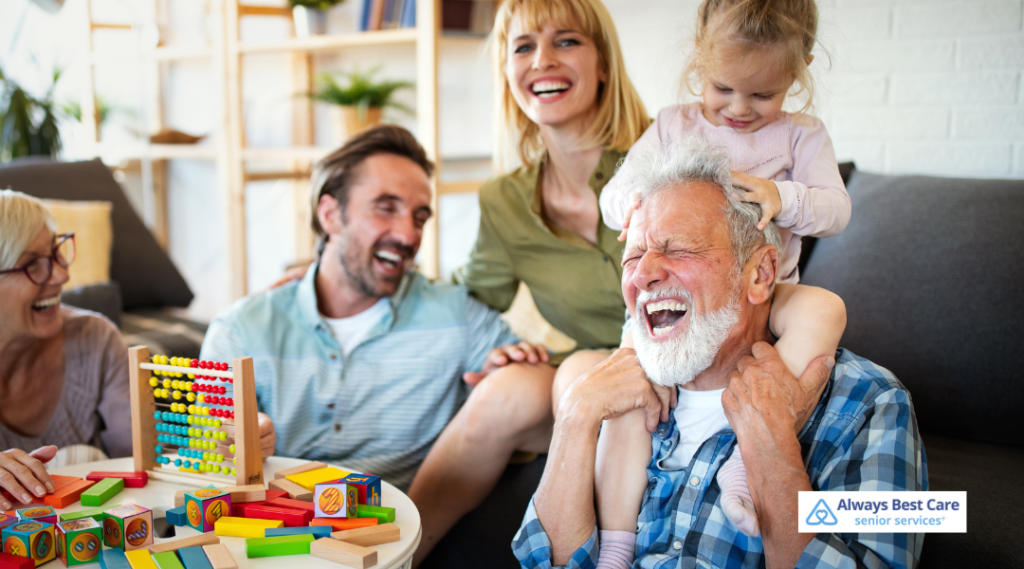 A joyful multi-generational family, including an older man, a small child, and other adults, laughing together in a bright living room with colorful toys on the table