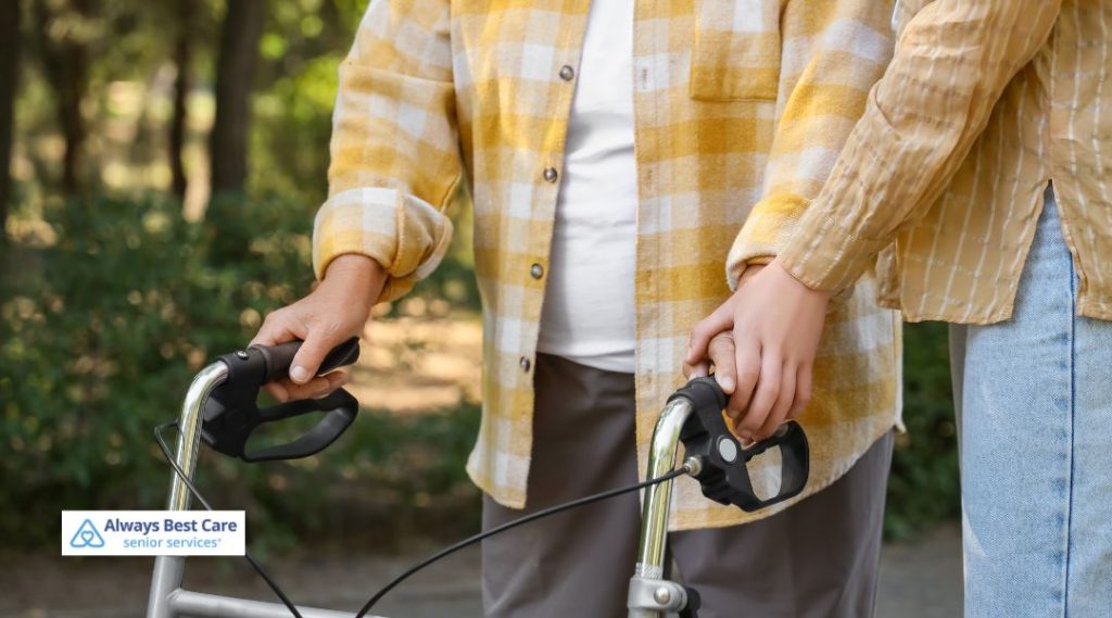A close-up of a caregiver assisting a senior with a walker outdoors. The caregiver gently holds the senior's hand, ensuring they have the support needed for safe walking, emphasizing care and companionship