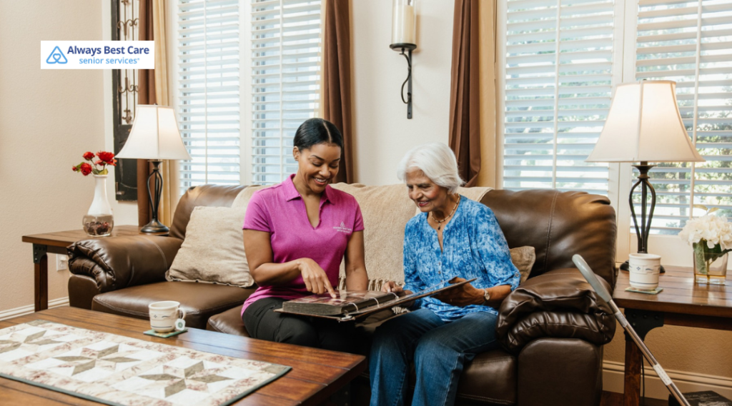 Caregiver and senior woman sitting together on a couch, looking through a photo album and sharing memories.