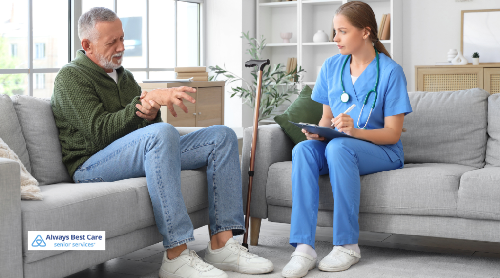 Caregiver in blue scrubs takes notes on a clipboard while an older man gestures during a conversation on a living room sofa; cane nearby; ‘Always Best Care senior services’ logo in the corner.
