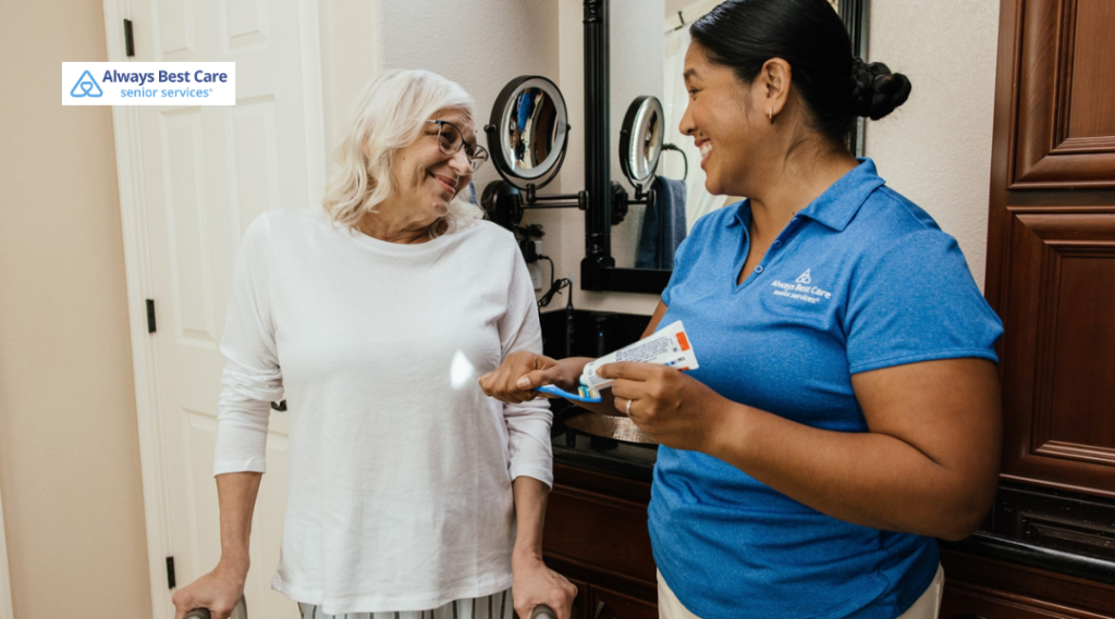 Home care aide in a blue polo smiles while helping an older woman with a toothbrush and toothpaste in a bathroom; ‘Always Best Care senior services’ logo in the corner.