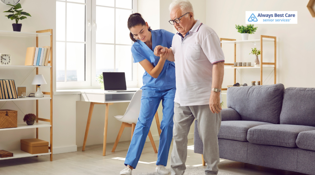 Smiling caregiver in blue scrubs holds a senior man's arm to steady him as he walks through his bright living room.