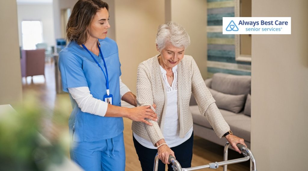 A caregiver in blue scrubs assists an elderly woman with a walker while offering support in a bright and spacious living room, showing the importance of mobility assistance.