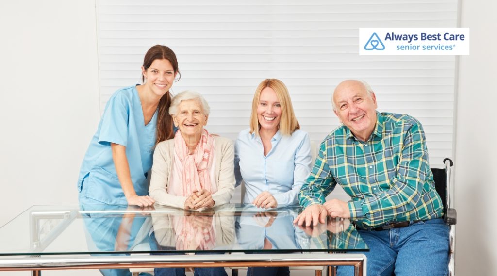 A caregiver in blue scrubs poses with an elderly woman and her family members around a glass table, sharing a joyful moment, highlighting compassionate care and family support.