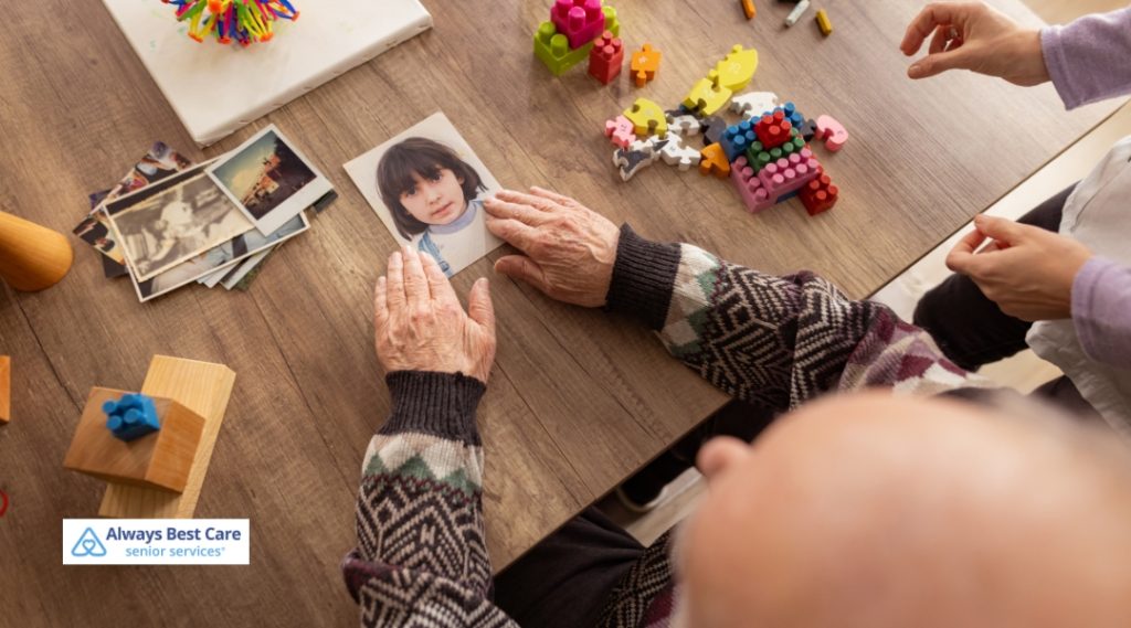 A senior examines an old photograph of a child on a wooden table scattered with nostalgic photos and colorful toys, engaging in memory care or reminiscence therapy.
