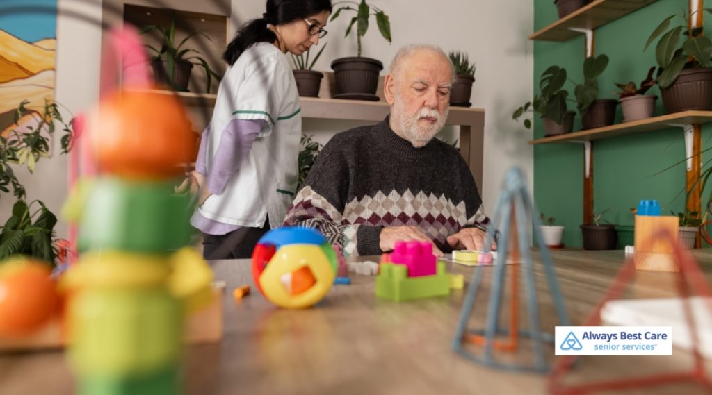 An elderly man concentrates on arranging colorful puzzle blocks at a table, with a caregiver nearby in a cozy, plant-filled room designed for therapeutic engagement.