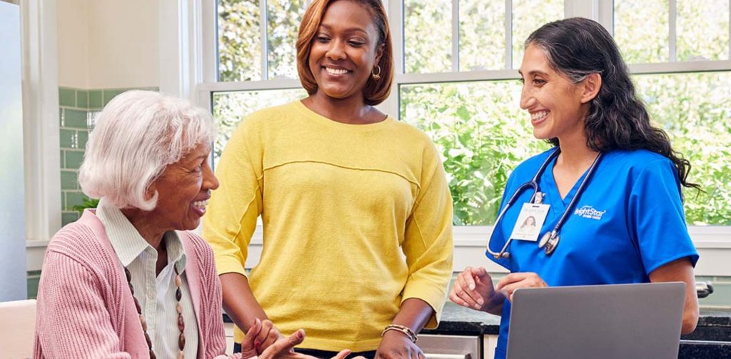 A BrightStar Care nurse in blue scrubs communicates with an older adult who is seated at a kitchen table. Another person stands close to them, wearing a yellow
