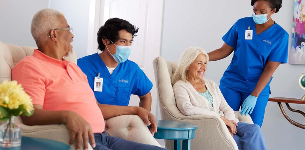 A professional caregiver in blue scrubs with a BrightStar Care logo sits beside two older adults in armchairs, engaged in a supportive conversation inside a cozy, well-lit room.