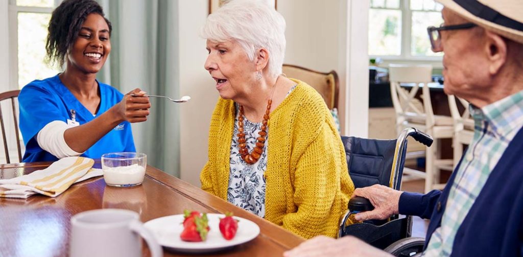 A caregiver in BrightStar Care blue scrubs assists a senior in a wheelchair, offering a spoonful of food during a meal at a dining table.