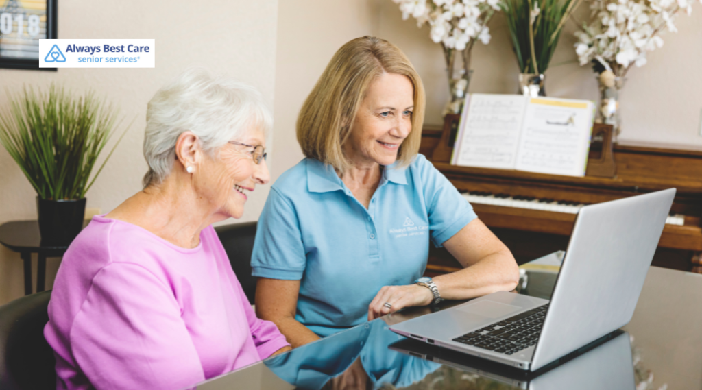 An elderly woman wearing glasses and a pink sweater sits at a table with a professional caregiver in a light blue polo shirt