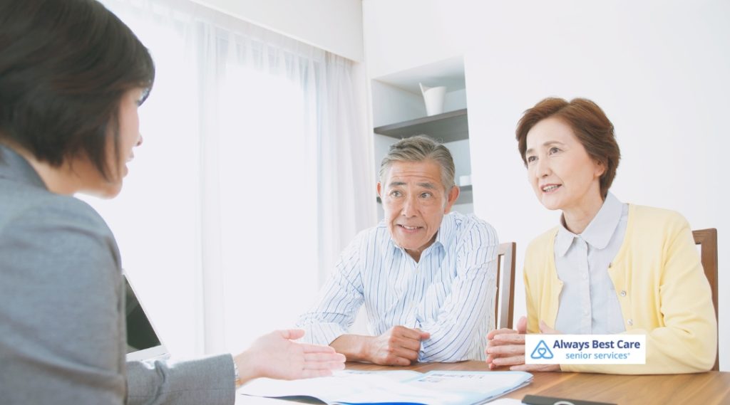 A female caregiver in light blue scrubs sits facing two older adults, speaking animatedly and referencing notes in an open binder. The Always Best Care Senior Services logo is present in the lower left corner.