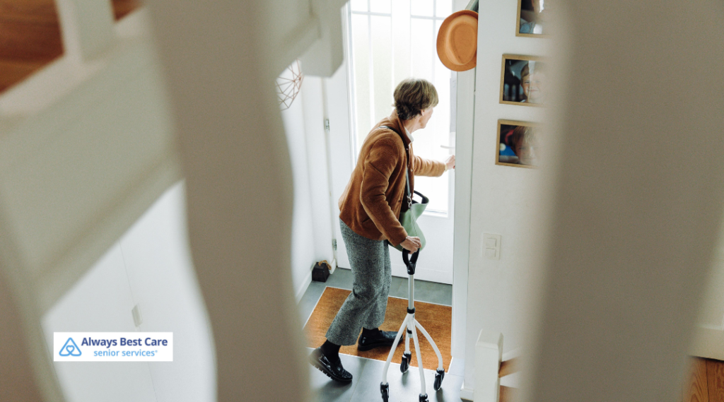 Older woman using a quad cane as she walks toward her front door, viewed through stair rails, illustrating safe independence at home with Always Best Care.