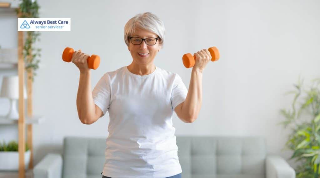 An older woman smiling and lifting a pair of orange dumbbells in a well-lit living room. She is dressed in a white shirt, focusing on her fitness and strength with a positive and energetic attitude.