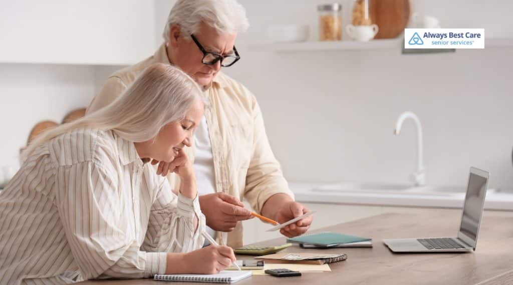 Older couple reviews paperwork and calculator at the kitchen table beside an open laptop.